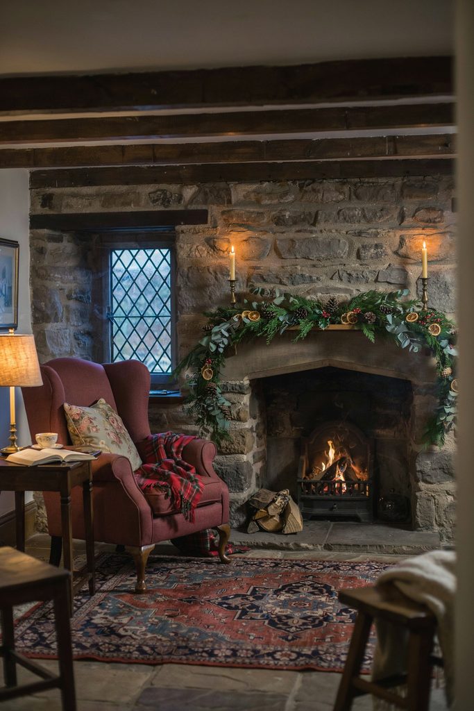 English cottage living room at Christmas with fresh cedar garland on a stone mantel, tartan throws, brass candlesticks, and a fire lit in the hearth
